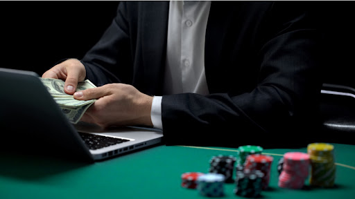 man sitting in front of laptop at poker table holding stack of cash